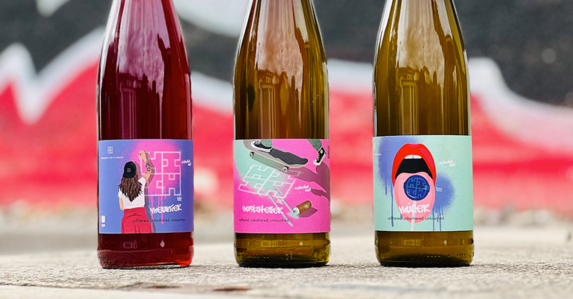 Drei Naturwein-Flaschen mit bunten Comic-Etiketten vor Graffiti-Hintergrund