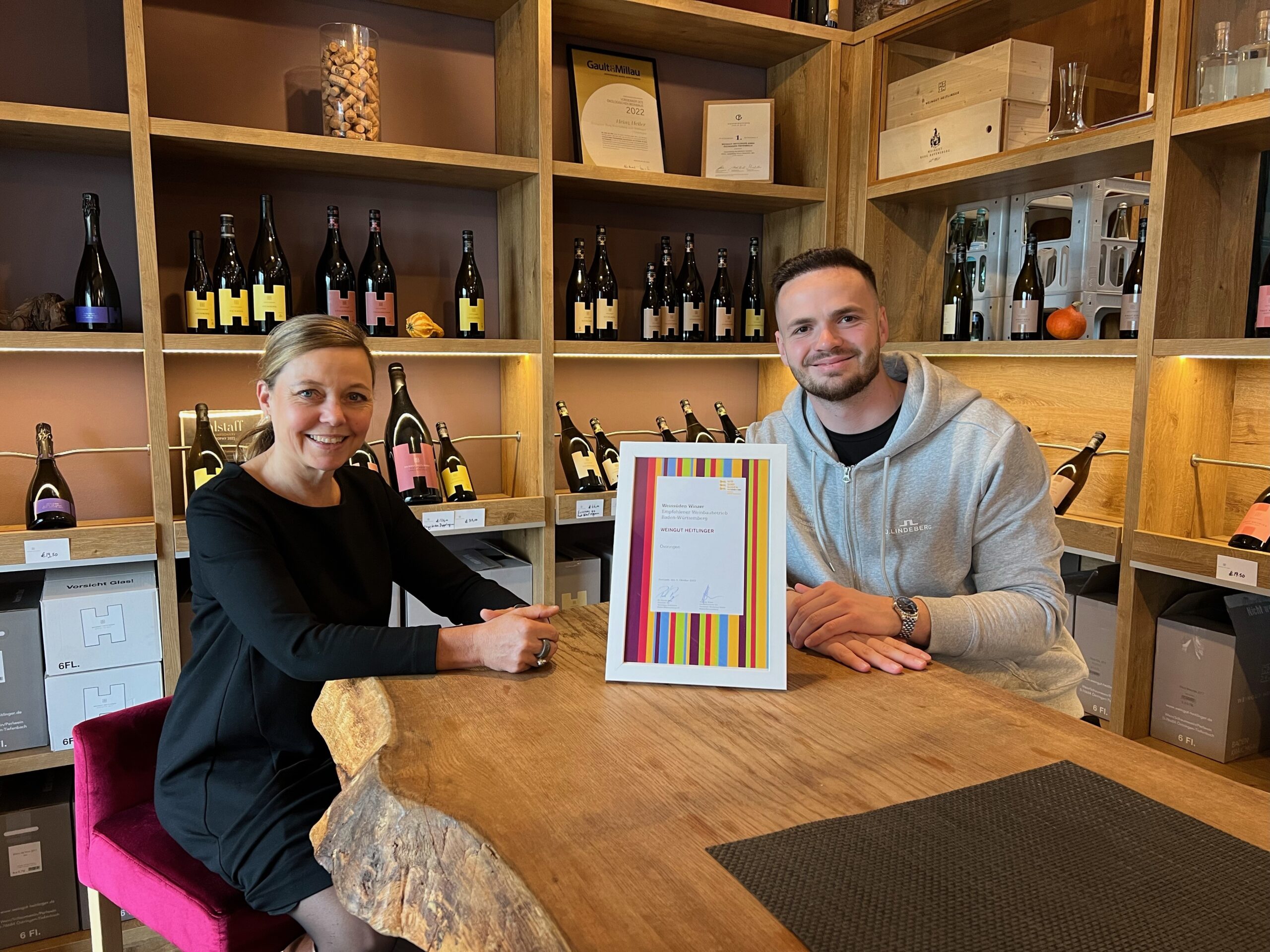 Frau und Mann mit Urkunde am Holztisch vor einem Weinregal