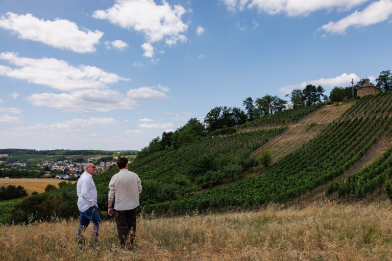 Zwei Männer auf einer Wiese vor einem steilen Weinberg mit Kapelle