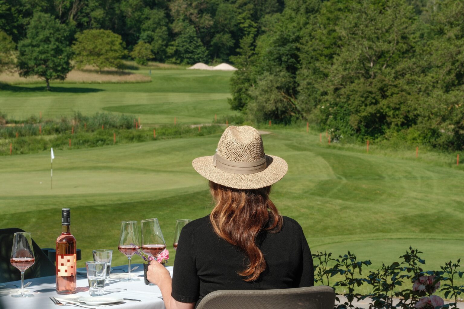 Frau mit Strohhut am gedeckten Tisch mit Roséwein, Blick auf den Golfplatz