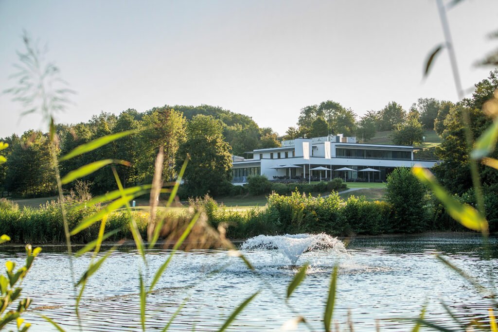 Neues Clubhaus hinter dem Teich mit Fontäne, Gräser im Vordergrund