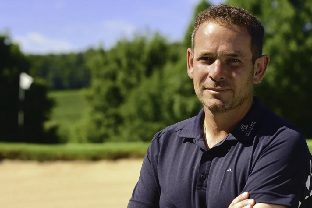 Warren Jacklin im dunklen Polo mit verschränkten Armen auf dem Golfplatz, Bunker und Fahne im Hintergrund
