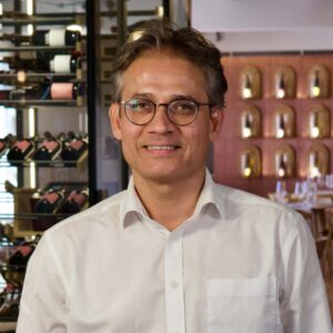 Gautam Nagpal mit Brille in weißem Hemd vor Weinregalen im Restaurant