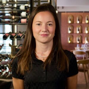 Geresa Arifi im schwarzen Polo vor Weinregalen im Restaurant