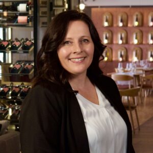 Jana Prestel in schwarzem Blazer und weißer Bluse im Restaurant, Weinregale im Hintergrund