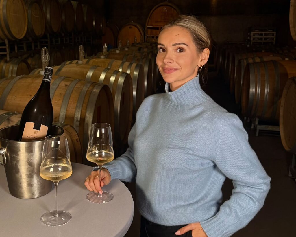 Marie mit Weißweinglas im Barriquekeller, daneben Sektkühler mit Flasche