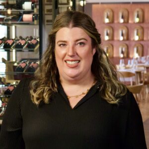 Svenja Bode lächelnd in schwarzer Bluse vor Weinregalen im Restaurant